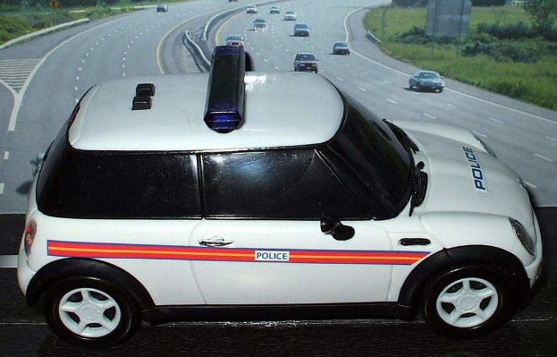 Weißer Polizeiwagen vor einer Reihe fahrender Fahrzeuge auf einer Straße mit Gras und Bäumen außerhalb der Stadt.