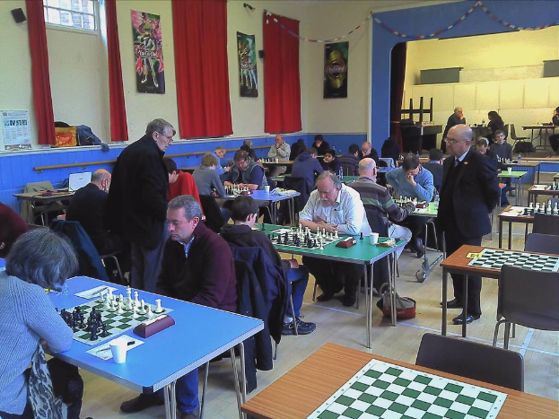 Ein großer Saal mit zwei Personen, die an einem Tisch Schach spielen, mit einem Kalender an der Wand, Vorhängen, Postern und einem Fenster im Hintergrund.