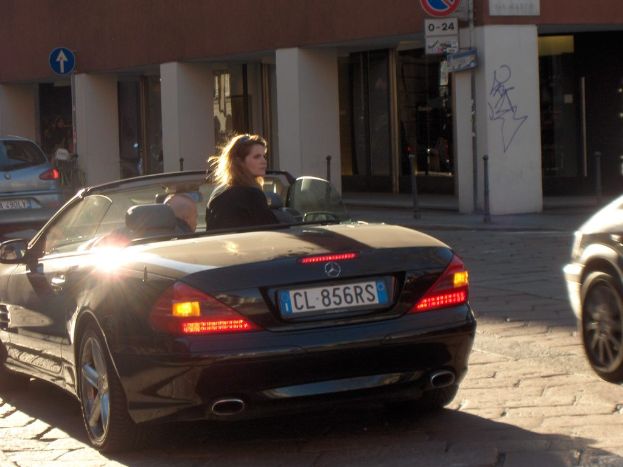 Eine Frau sitzt nachts in einem Auto auf einer Straße, mit Gebäuden, Säulen, einem Schild und anderen Fahrzeugen im Hintergrund.