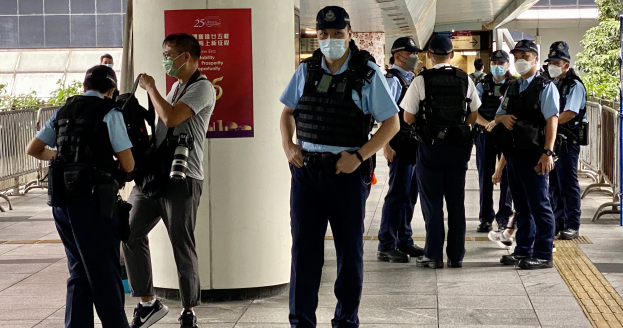 Eine Gruppe von Polizisten in Uniform und Mund-Nasen-Schutz steht vor einem Gebäude, einige halten Kameras, mit einem Banner an einer Säule, Geländern, Pflanzen, Bäumen und Deckenlampen im Hintergrund.