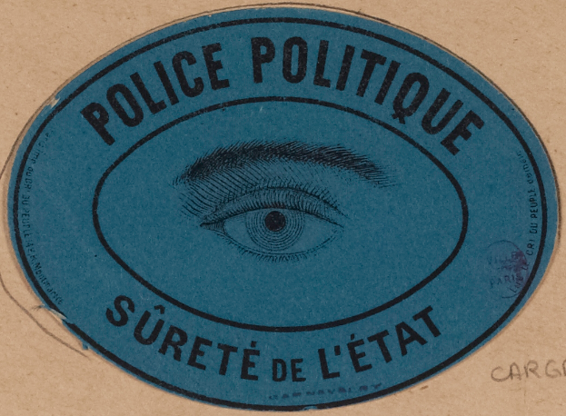 Ein brauner Karton mit einem blauen Aufkleber, auf dem "Polizei Politique Surete de L'État" und ein Auge in der Mitte steht.