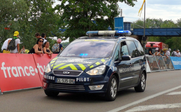 Polizeiauto fährt eine Straße entlang neben einer Menschenmenge, mit einem Banner auf der linken Seite, Geländern mit Bannern dahinter, Bäumen, einer Brücke, einer Fahne auf einem Mast und einem bewölkten Himmel im Hintergrund.