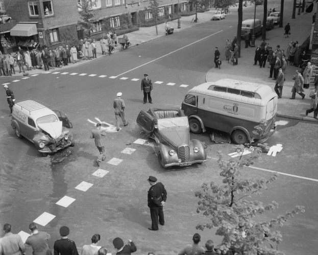 Schwarz-weiß-Szene eines Autounfalls an einer Straßenkreuzung mit einem Van, anderen Fahrzeugen und Menschen, die sich drum herum versammelt haben, mit Gebäuden, Bäumen, Pfählen und Texttafeln im Hintergrund.