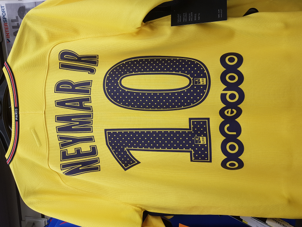 Ein gelbes Fußballtrikot mit der Nummer 10, Teil des Heimtrikots von 2018-19 von PSG, das Neymar Jr. gehört, mit einem schwarzen Etikett und Papieren und anderen Gegenständen im Hintergrund.