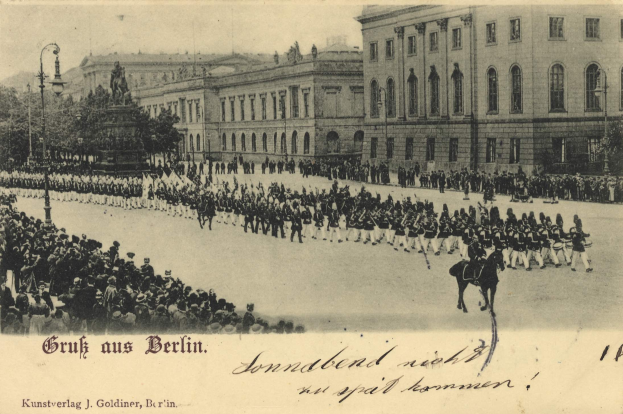 Ein altes Schwarz-Weiß-Foto eines Umzugs in Berlin, Deutschland, mit Menschen auf dem Boden und auf Pferden, einer Statue, Straßenlaternen, Bäumen, Gebäuden und einem bewölkten Himmel, mit Text unten.