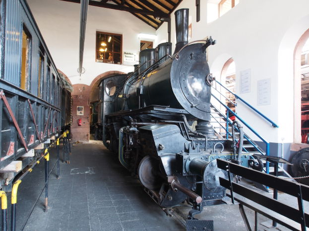 Alte Dampflokomotive in Museumsgebäude mit Informationsschildern, Geländern und Beleuchtung ausgestellt.