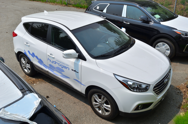 Drei Autos nebeneinander auf einem Parkplatz geparkt, mit einem Maschendrahtzaun und Gras im Hintergrund, ein Auto zeigt den Text 'Wasserstoff-getriebenes Fahrzeug'.