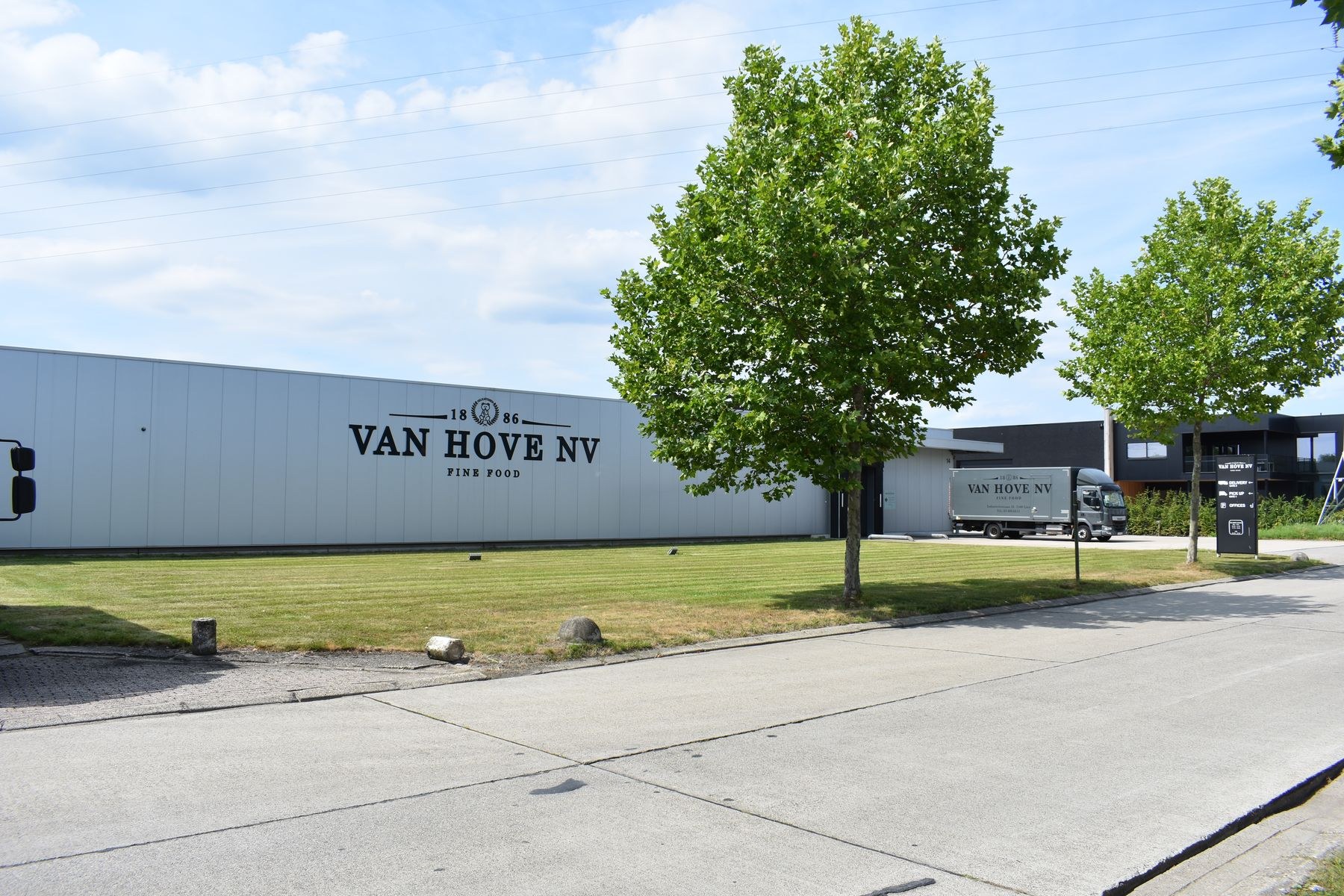 Van Hove NV Einrichtung in den Niederlanden mit Fahrzeugen auf der Straße, Gras, Bäumen, Gebäuden, Brettern, Pfählen und anderen Gegenständen im Vordergrund und einem bewölkten Himmel im Hintergrund.