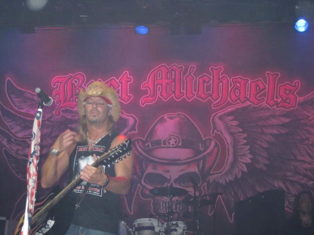 Eine Musikkonzert-Szene mit einem Gitarristen links neben einem Mikrofon, einer Person in der rechten unteren Ecke, einem Banner im Hintergrund und Drogen in der Mitte unten, alles unter Bühnenbeleuchtung von oben.