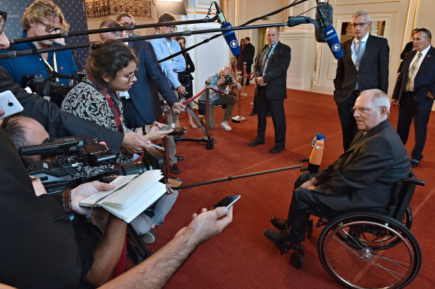 Ein Mann im Rollstuhl spricht mit Reportern, die Kameras, Handys und Mikrofone halten, vor einem Banner, Lichtern und einer Wand.