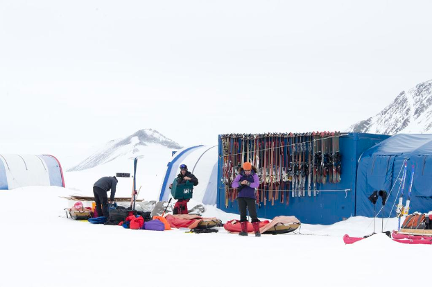 Drei Personen auf einer schneebedeckten Landschaft mit Taschen und Zelten, Skier lehnen an den Zelten, mit schneebedeckten Hügeln und einem klaren Himmel im Hintergrund.
