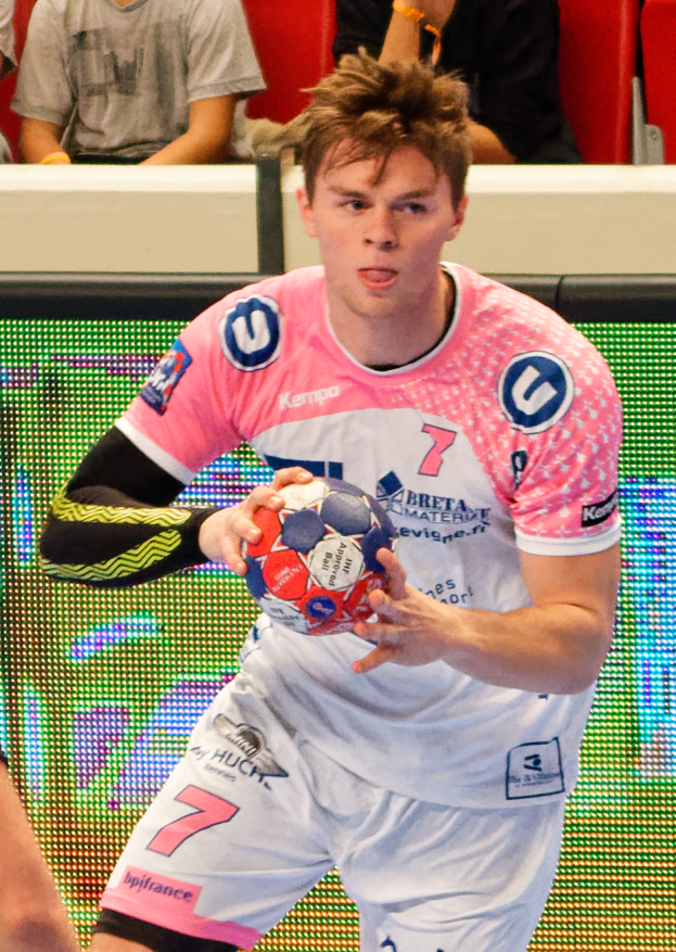 Ein Mann in einem pinken Hemd hält einen Fußball, mit der Hand eines anderen Menschen auf der linken Seite und einigen Menschen im Hintergrund auf Stühlen sitzend.