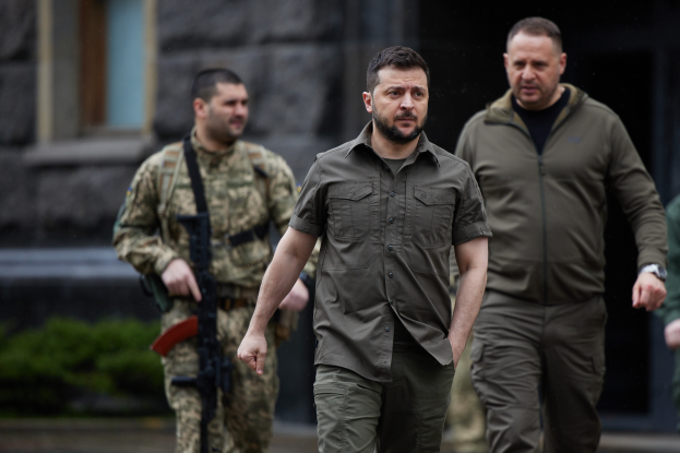 Ein Mann in militärischer Uniform, identifiziert als der ehemalige ukrainische Präsident Viktor Janukowytsch, wird von zwei bewaffneten Männern in militärischer Uniform vor einem Gebäude mit Pflanzen im Hintergrund eskortiert.