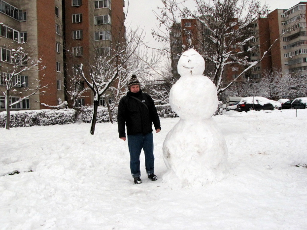 Ein Mann in einer schwarzen Jacke und blauen Jeans steht neben einem Schneemann, mit schneebedeckten Sträuchern, Bäumen, Autos und Gebäuden im Hintergrund.