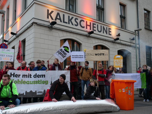 Gruppe von Menschen mit Protestschildern und Plakaten vor einem Gebäude, mit zwei Personen im Vordergrund und einem Müllimer rechts, vor dem Hintergrund von Gebäuden mit Fenstern und Schildern in Deutschland.