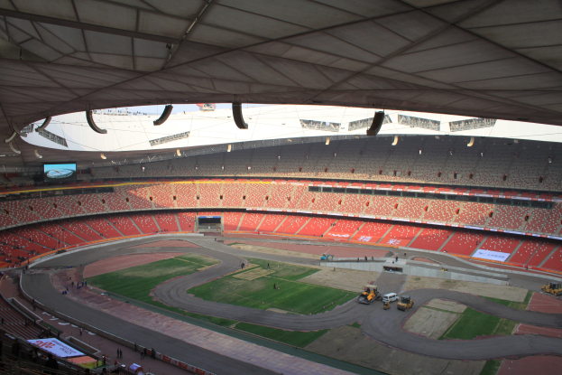 Ein großes Stadion voller Menschen, umgeben von Fahrzeugen auf einer Straße, Gras, Texttafeln, Geländern, einem Bildschirm und Deckenleuchten.