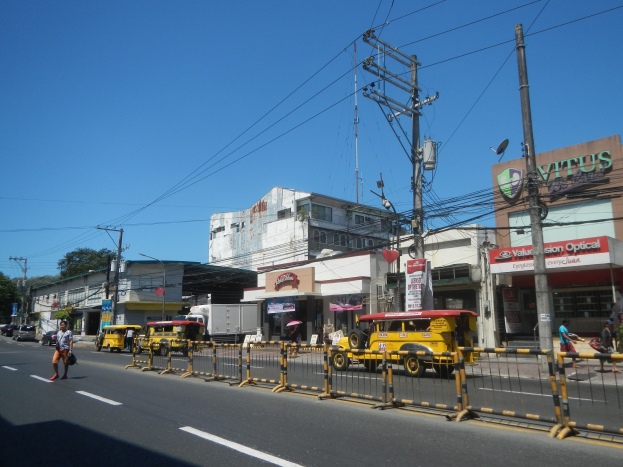 Eine belebte Stadtstraße mit hohen Gebäuden, mehreren Fahrzeugen, Fußgängern auf dem Gehweg, Geländern, Strommasten mit Drähten, Gebäudeschildern, Bäumen und einem klaren blauen Himmel.