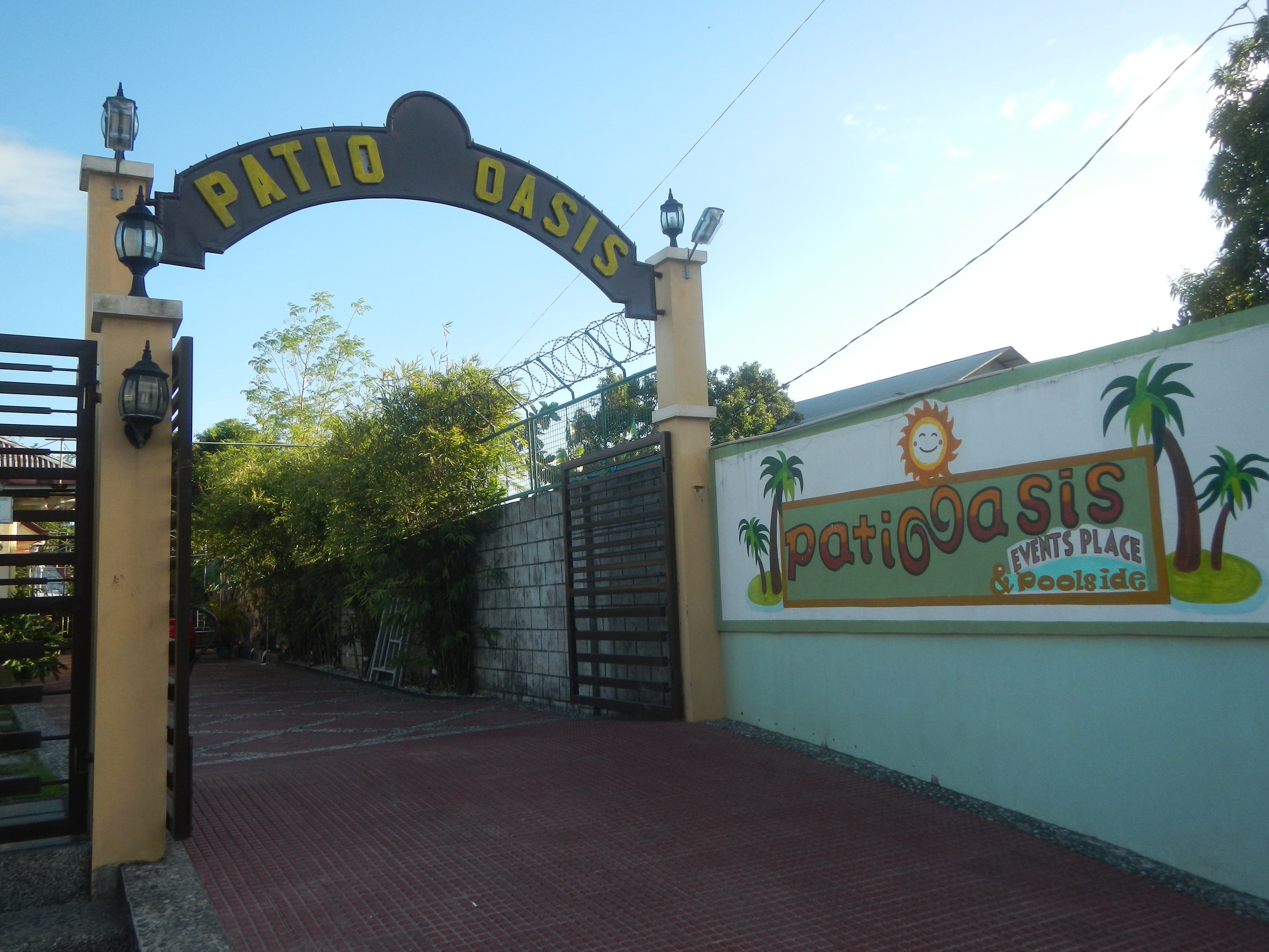 Eintritt zum Patio Oasis Events Place & Poolside mit einem Bogen mit Text, einer bemalten Wand, einem Tor, Wandbeleuchtung, Bäumen und einem bewölkten Himmel.