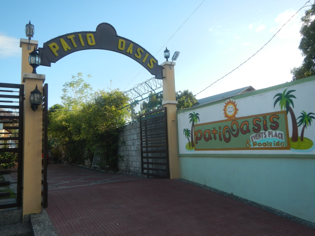 Eintritt zum Patio Oasis Events Place & Poolside mit einem Bogen mit Text, einer bemalten Wand, einem Tor, Wandbeleuchtung, Bäumen und einem bewölkten Himmel.