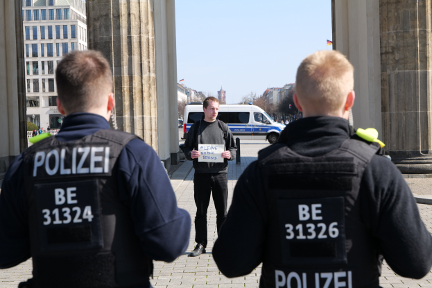 Drei Polizeibeamte stehen vor einem Gebäude, ein Mann in der Mitte hält ein Papier, flankiert von Säulen, während Fahrzeuge, Menschen, Bäume und Gebäude den Hintergrund und den Himmel ausfüllen.