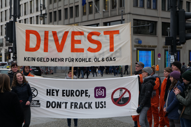 Eine Gruppe von Menschen hält ein Banner mit der Aufschrift "Divest Hey Europe, Don't Frack Us" auf einer Straße mit Gebäuden, Straßeninfrastruktur, Fahrzeugen, Bäumen und einem klaren Himmel im Hintergrund.