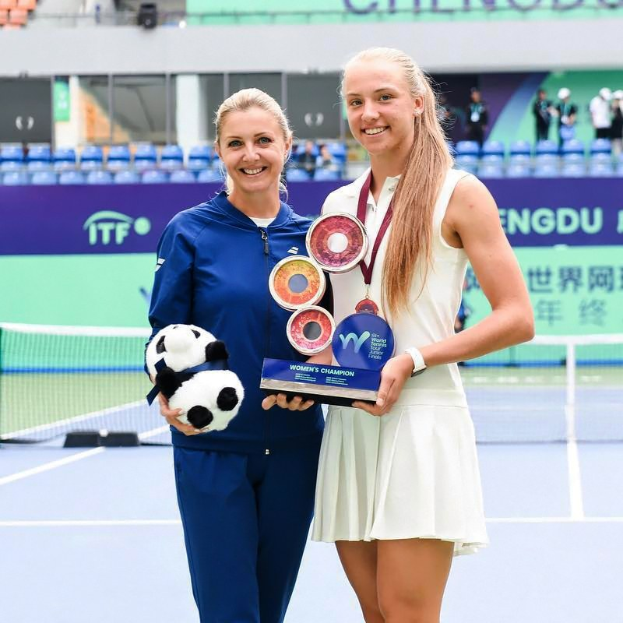 Zwei Frauen in Tennisbekleidung stehen auf einem Court und halten Trophäen, eine in einem blauen Kleid, die eine Kugel hält, mit Netz und Zuschauern im Hintergrund.