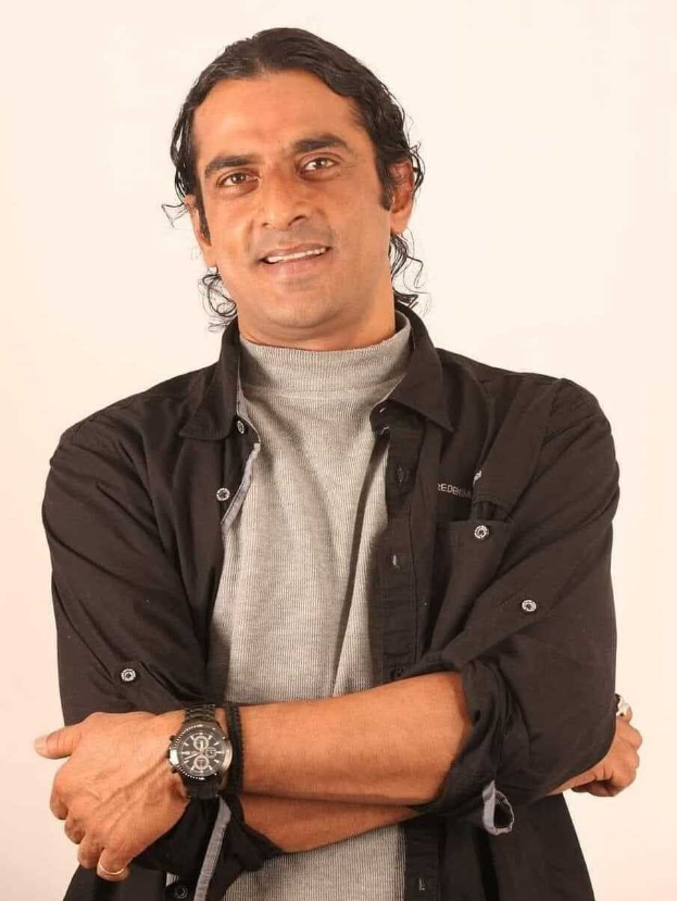 Ein Mann in einem schwarzen und aschfarbenen Kleid mit überschlagenen Armen, der eine Uhr tr├Ągt und gegen einen wei├čen Hintergrund steht, der nachdenklich erscheint.