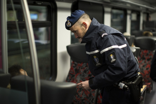 Ein Polizeibeamter in Uniform und Mütze steht in einem Zug und schaut auf sein Handy, umgeben von sitzenden Passagieren und einer weiteren Person, die rechts steht.
