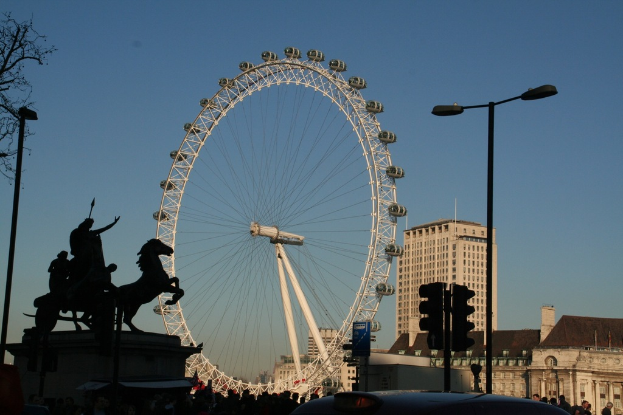 Gebäude und Masten auf der rechten Seite, ein großes Riesenrad in der Mitte und eine Skulptur auf der linken Seite.