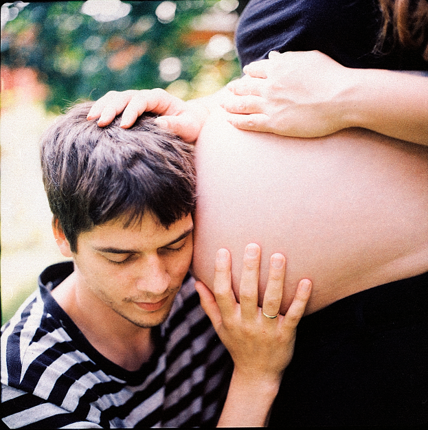 Ein Mann und eine Frau stehen nebeneinander, wobei der Mann zärtlich den Bauch der Frau berührt und auf ihr ungeborenes Kind schaut.