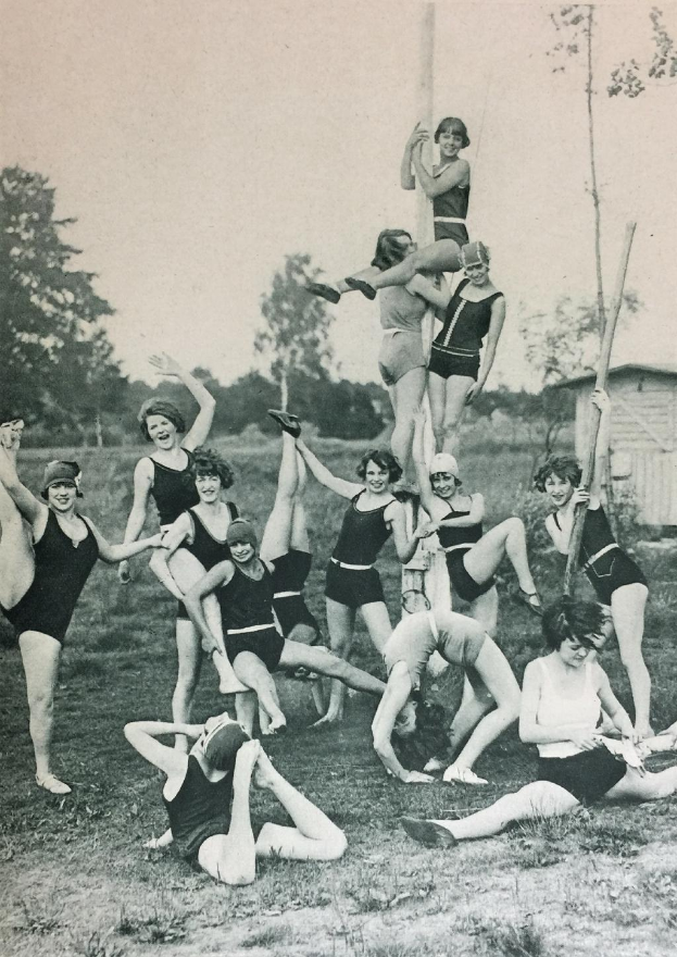 Eine Gruppe Frauen in Badeanzügen posiert für ein Foto auf einer Wiese, umgeben von Bäumen und einem Haus im Hintergrund, schwarz-weiß, 1920er Jahre.