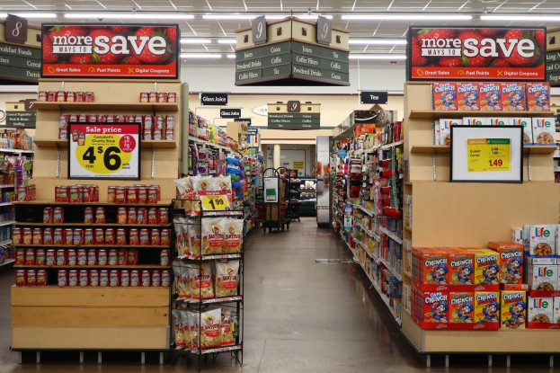 Ein Kiosk-Innenraum mit Regalen und Boden voller verschiedener Lebensmittel, beleuchtet von Deckenlampen und mit Informationsschildern; im Hintergrund ist eine Person zu sehen.