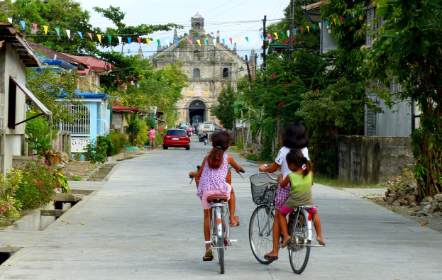 Drei Kinder fahren Fahrräder auf einer von Bäumen gesäumten Straße mit Gebäuden auf beiden Seiten, ein Auto und ein Gebäude im Hintergrund und Himmel darüber.