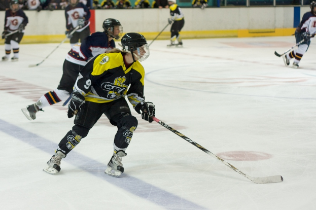 Menschen beim Eisstockschießen mit Eishockeyschlägern und Helmen, mit anderen und einer Wand im Hintergrund.