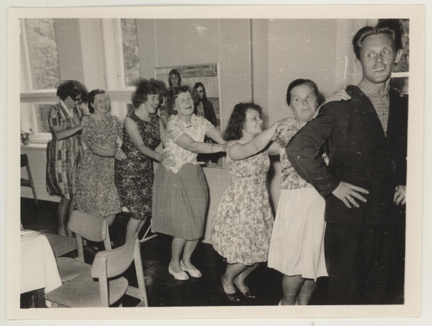 Gruppe von Menschen, die in einem Raum im Stil der 1950er Jahre tanzen, mit Stühlen, einem Tisch mit wei├čer Decke, einem Foto auf der Wand und Fenstern im Hintergrund.
