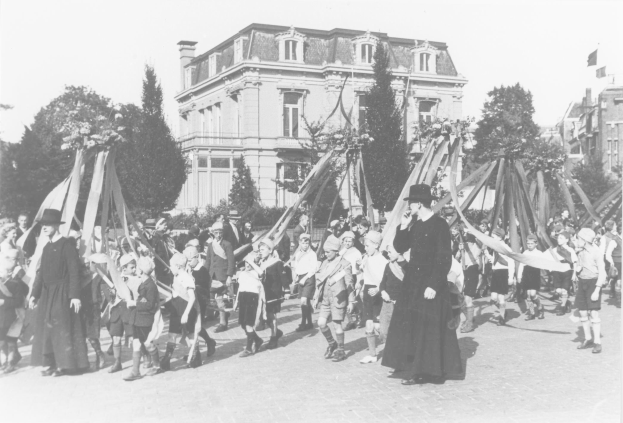 Schwarüßes Bild einer Gruppe von Menschen, die eine Straße entlanggehen, einige halten Fahnen, mit Hüten, Bäumen, Gebäuden und einem sichtbaren Himmel im Hintergrund, mit einem Maypole, das auf ein Mai-Fest hinweist.