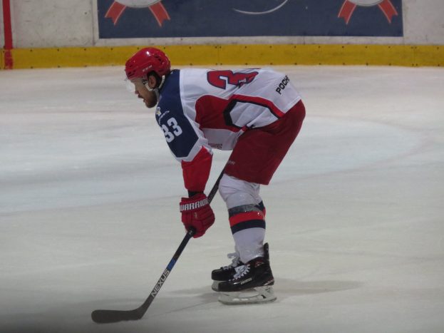 Hockeyspieler in voller Ausrüstung auf dem Eis mit einem Banner im Hintergrund, das Text und Logos enthält.