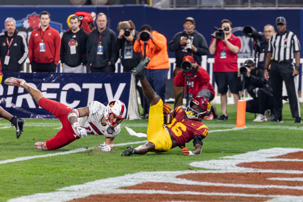 Ein Football-Spieler taucht in die Endzone ein, um einen Touchdown zu erzielen, mit einer Gruppe von Menschen am Boden, einige halten Kameras, und ein Banner mit Text im Vordergrund, während Geländer, Pfosten und andere Gegenstände im Hintergrund sichtbar sind.