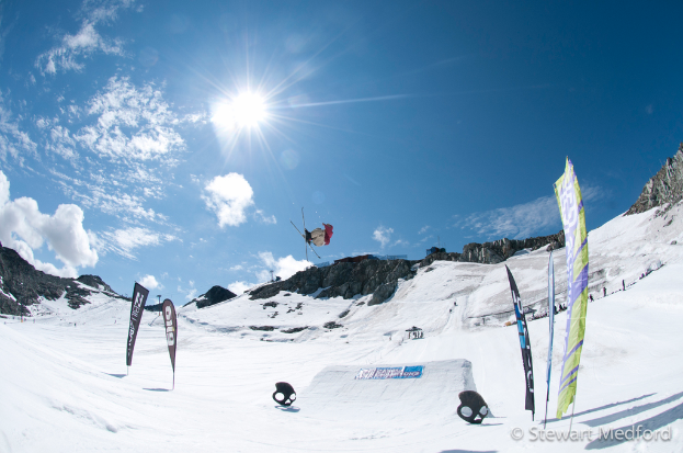 Eine Person in der Luft auf Skiern mit schneebedeckten Bergen im Hintergrund.