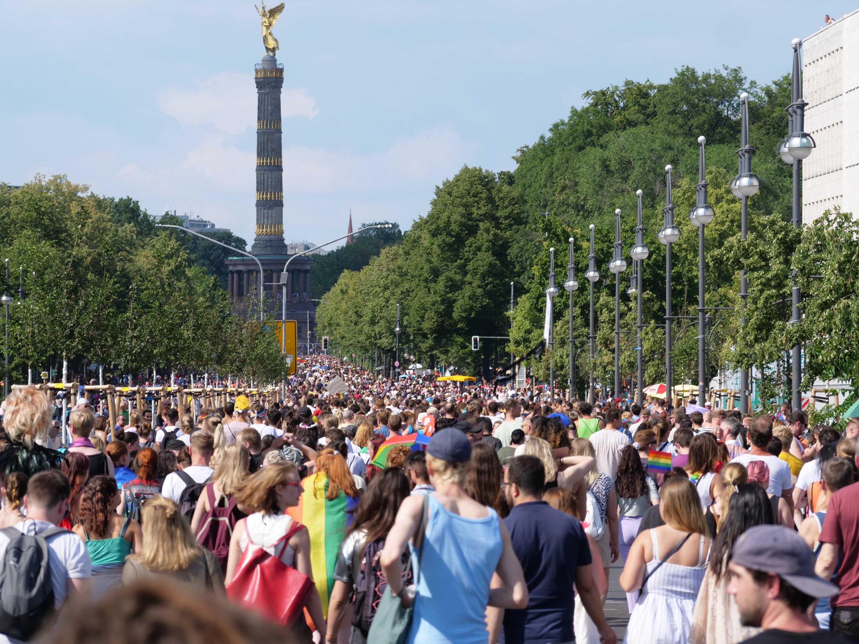 Eine große Menschenmenge mit Taschen geht eine Straße in Berlin, Deutschland, entlang, mit Laternen am Weg, Bäumen und einem Turm mit einer Statue im Hintergrund und umliegenden Gebäuden unter einem bewölkten Himmel.