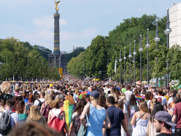Eine große Menschenmenge mit Taschen geht eine Straße in Berlin, Deutschland, entlang, mit Laternen am Weg, Bäumen und einem Turm mit einer Statue im Hintergrund und umliegenden Gebäuden unter einem bewölkten Himmel.