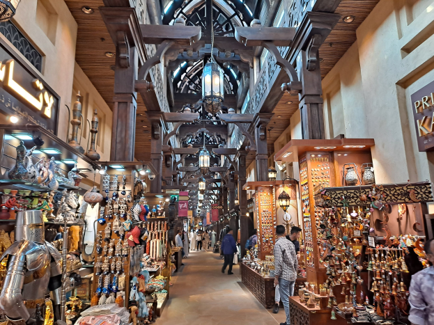 Ein geschäftiges Dubai-Bazar mit Menschen, die zwischen Regalen voller Waren hindurchgehen, beleuchtet von Deckenlampen und mit Texttafeln an den Wänden.