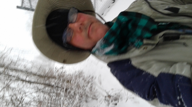 Eine Person in einer Jacke und einem Hut steht mit Schnee dahinter und Bäumen in der unteren linken Ecke.