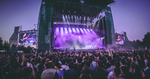 Eine große Menschenmenge auf einem Musikfestival, mit einer Bühne in der Mitte, auf der Musiker Instrumente spielen, beleuchtet von Lichtern und umgeben von Bildschirmen, und einem Gebäude, Bäumen und einem klaren blauen Himmel im Hintergrund.