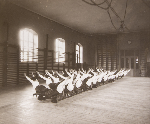 Gruppe von Menschen, die Pilates-Übungen in einem Fitnessstudio mit einer Uhr an der Wand machen.