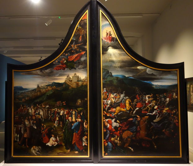 Ein großes Triptychon-Gemälde mit detaillierten Elementen und leuchtenden Farben wird in einem Museum ausgestellt, mit einem Foto Rahmen an der benachbarten Wand und Deckenlampen darüber.