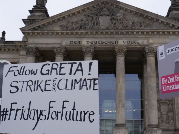 Ein große Gebäude mit Säulen und Skulpturen, das ein Schild mit der Aufschrift "Follow Greta Strike for Climate Fridays for Future" trägt, vor einem klaren blauen Himmel.