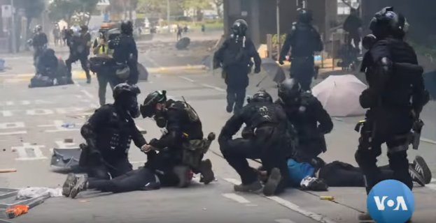 Eine Gruppe von Polizeibeamten in Einsatzausrüstung umringt einen Mann, der auf dem Boden liegt, mit Bäumen, Masten, Gebäuden und anderen Gegenständen im Hintergrund.
