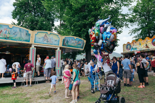 Eine Menschenmenge, einige mit Kinderwagen, versammelte sich um ein bunt beleuchtetes Karussell auf einem Volksfest, mit Bäumen und einem bewölktem Himmel im Hintergrund.