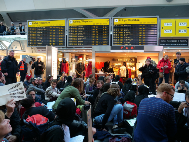 Eine große Gruppe von Menschen sitzt und steht in einem Flughafen während einer Demonstration, mit Informationsschildern, Schaufensterpuppen in Kleidern und Deckenbeleuchtung im Hintergrund.
